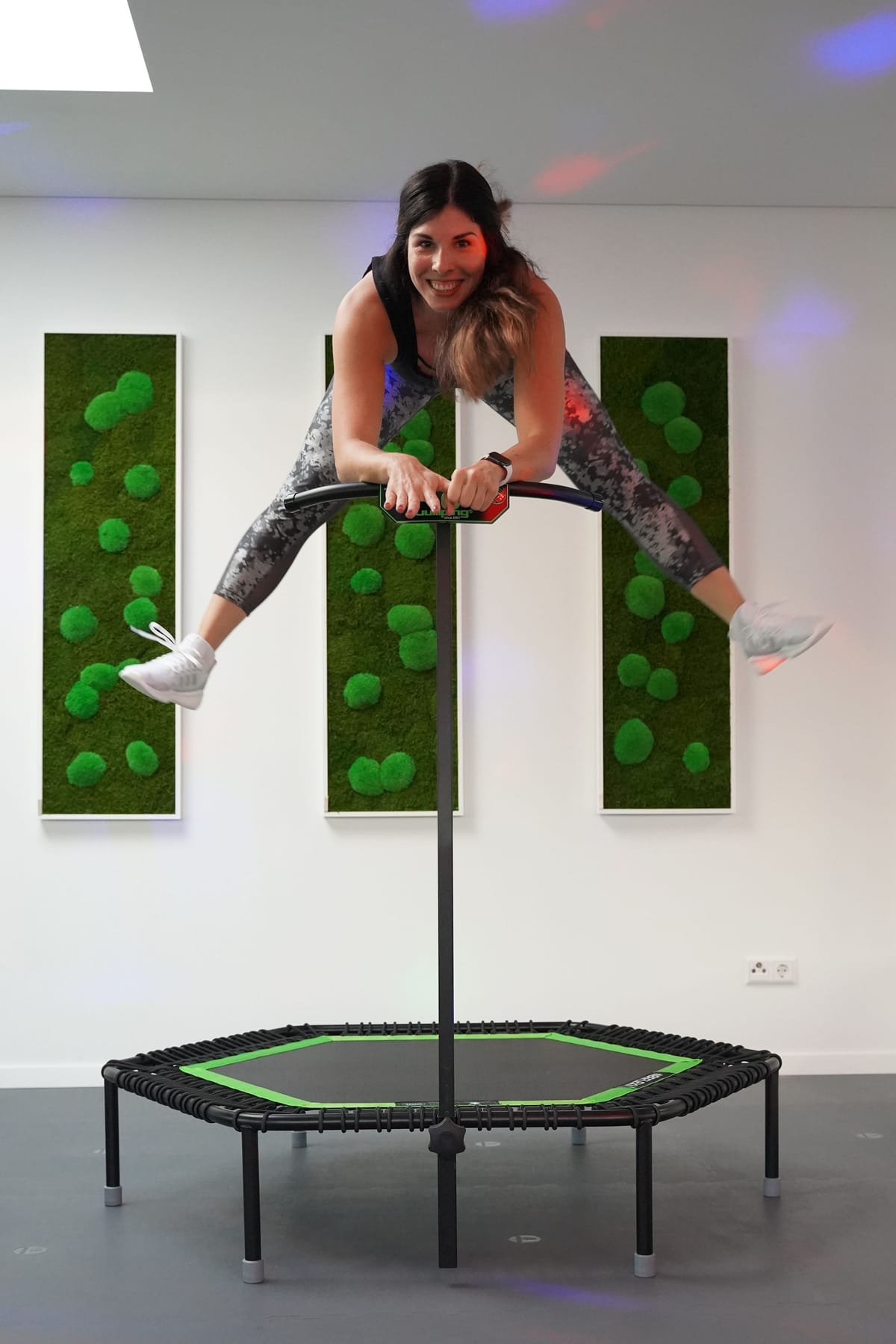 Trainerin im Sprung auf einem Jumping-Trampolin, dahinter die begrünten Wände des Studios